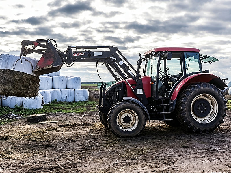 Red farm tractor fitted with front loader moving wrapped hay bales in farm field
