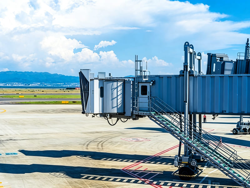 Steel passenger boarding bridge with access stairs on airport tarmac, blue sky with white clouds, distant mountains and runway in background
