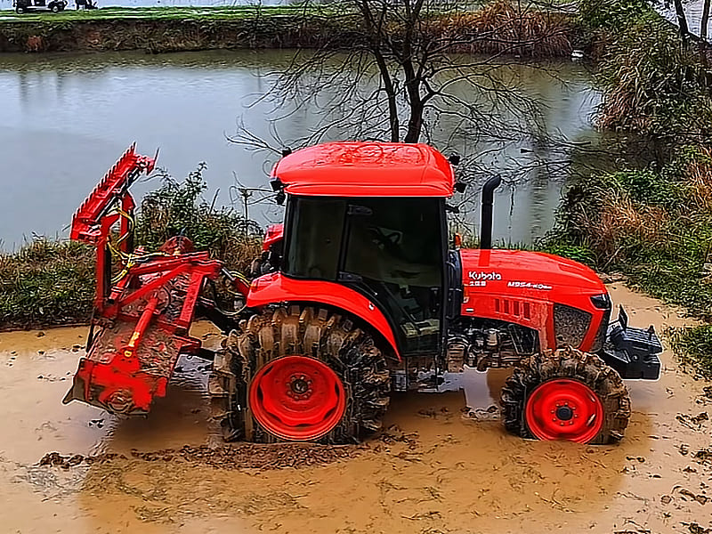 Red Kubota tractor equipped with rotary tiller, working in muddy farmland by a pond, tires covered with mud, outdoor agricultural scene