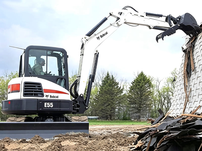 White Bobcat E55 compact tracked excavator performing building demolition, bucket tearing down wall structure, operator in cabin, outdoor construction site