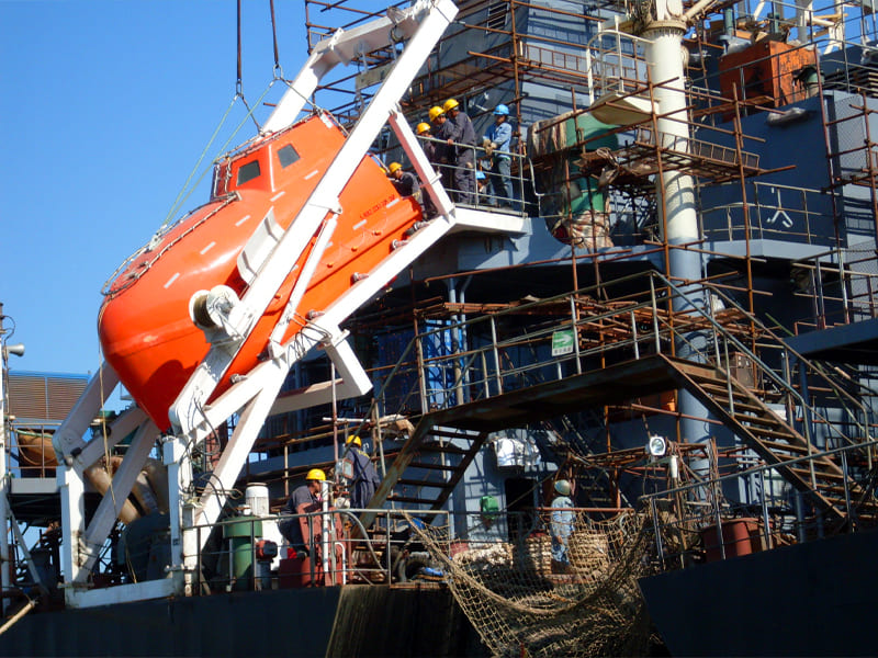 Orange enclosed marine lifeboat with white davit system, shipyard installation scene, workers with safety helmets, scaffolding on vessel deck