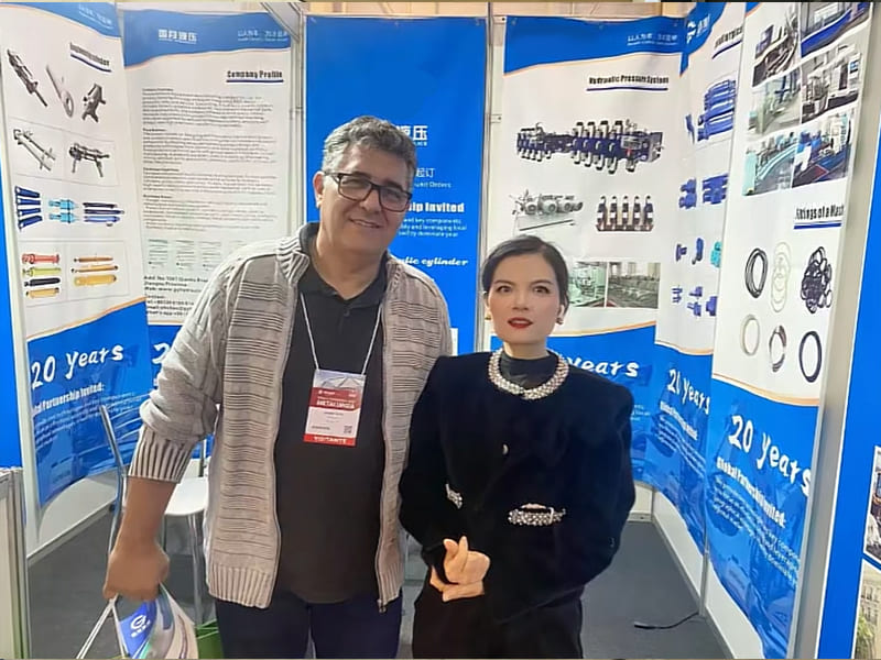 Female sales representative and foreign client take photo at hydraulic cylinder booth, display banners show hydraulic cylinders, sealing parts and company introduction