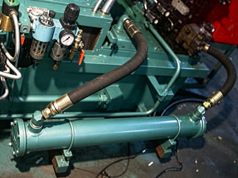 Industrial shell‑and‑tube hydraulic oil heat exchanger connected with high‑pressure rubber hoses on workshop machinery