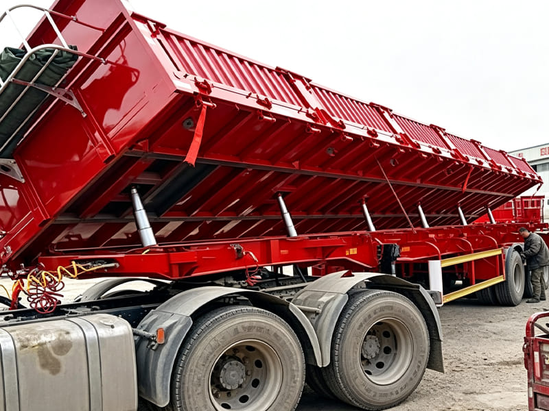 Red side‑tipping dump semi‑trailer lifted by several hydraulic cylinders, workshop yard