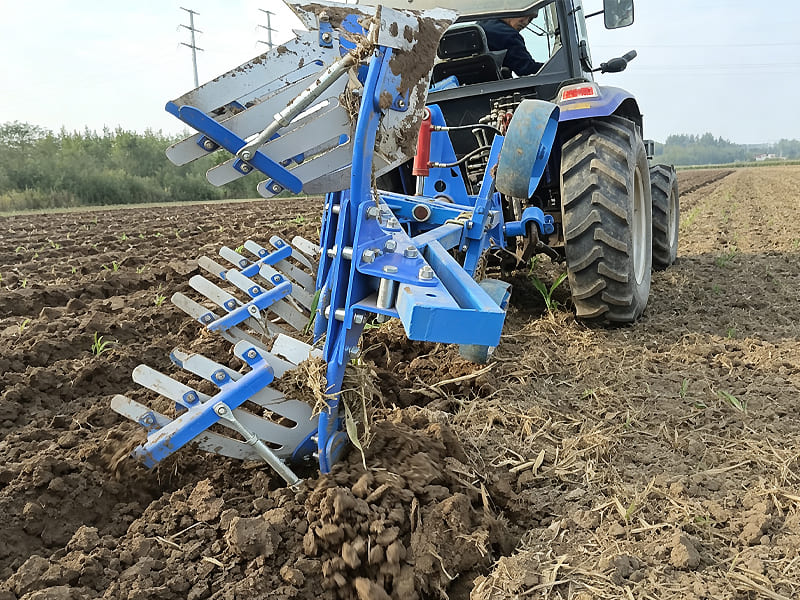 Blue reversible plough mounted on tractor, tilling soil in farm field