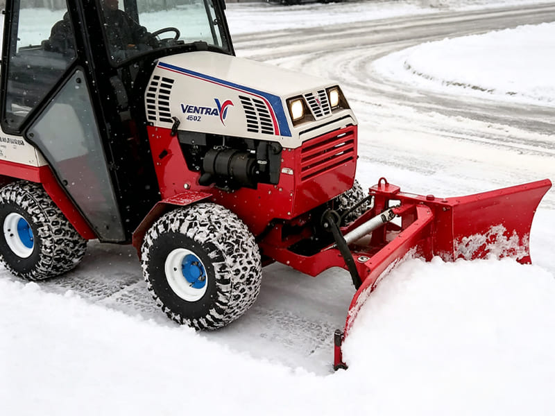 Red compact snow removal tractor with front snowplow working on snow ground