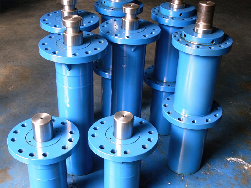 Multiple blue heavy hydraulic cylinders with large flanges stacked in factory warehouse