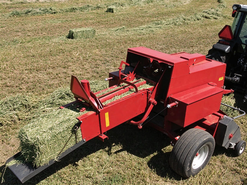 Red square hay baler attached to tractor, baling dried alfalfa grass in farm field, finished hay bales on ground