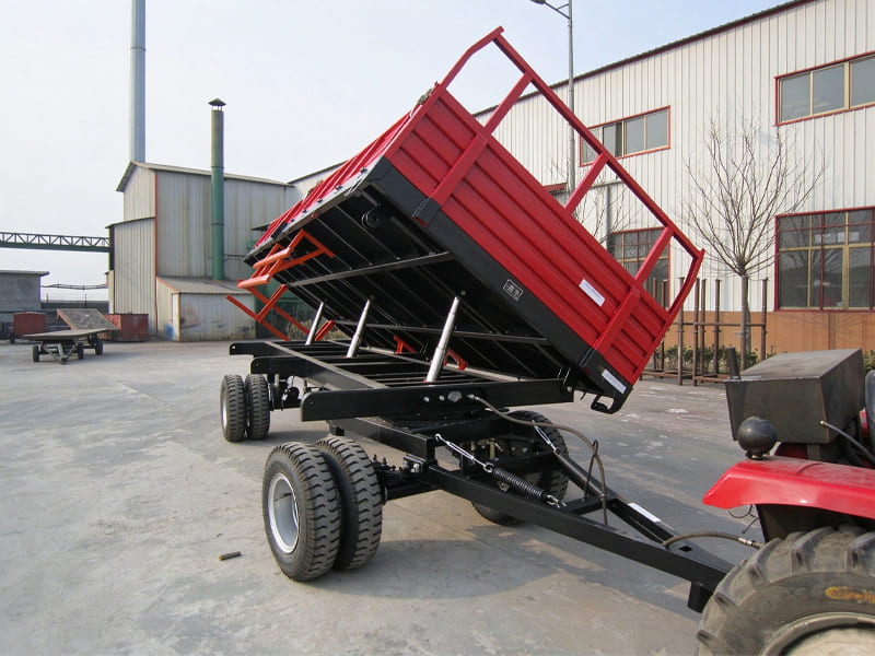 Red hydraulic dump farm trailer hitched to tractor, multi-axle tipping agricultural trailer with lifting hydraulic cylinders in factory yard