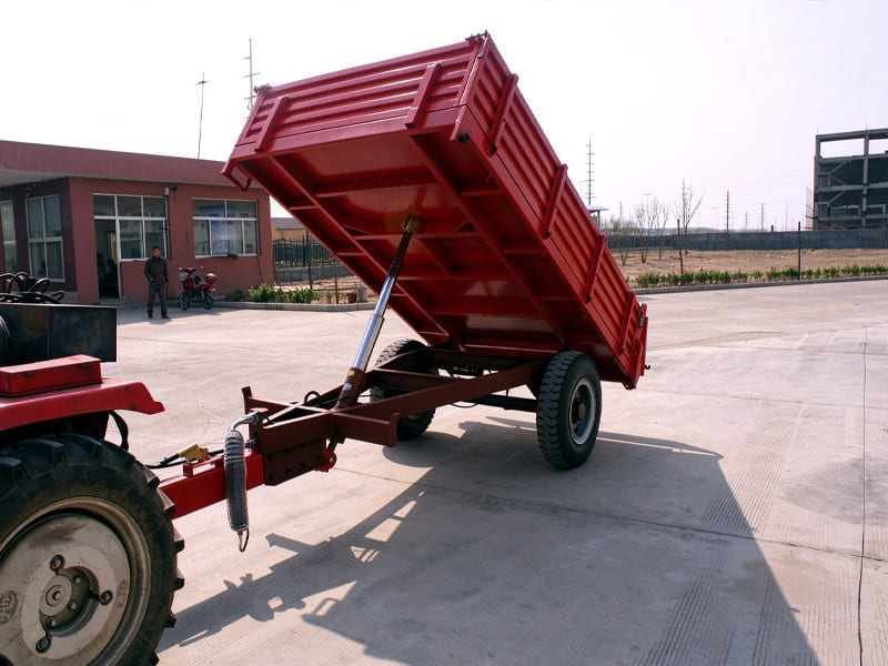 Red single axle agricultural tipping trailer, connected with tractor, hydraulic cylinder lifting the cargo bin